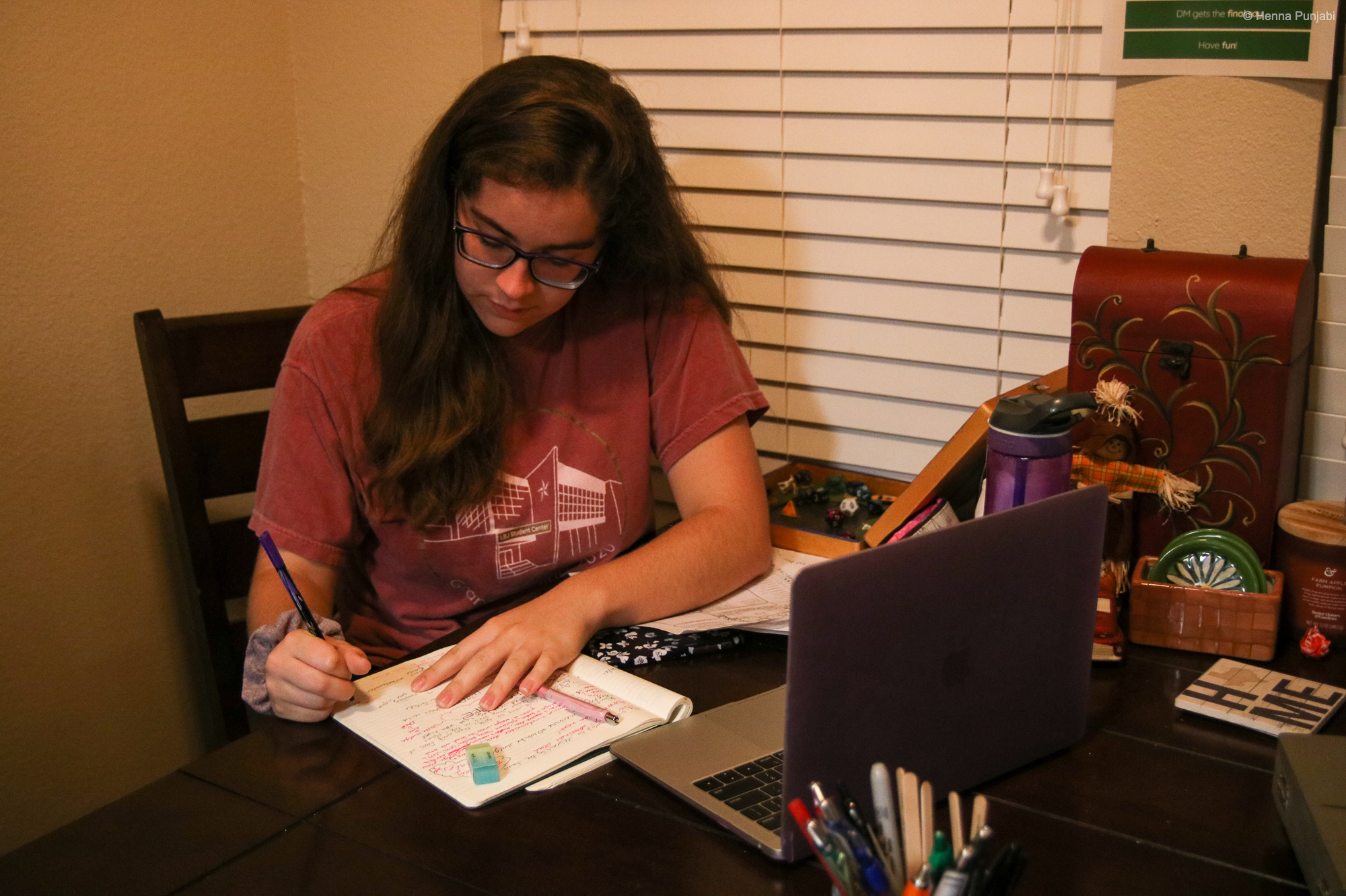 girl sitting at a table with a computer in front of her, taking notes in her notebook